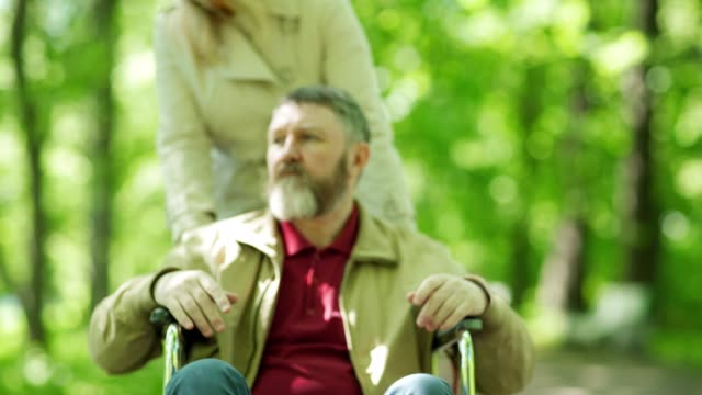 Zoom-in-of-female-caregiver-pushing-man-in-wheelchair-towards-camera-in-park.-Tired-senior-man-looking-at-camera