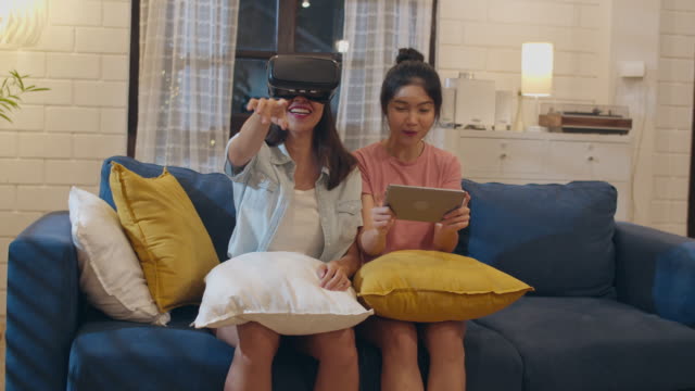 Lesbianas-mujeres-lgbt-pareja-usando-tableta-en-casa,-mujer-asiática-se-siente-feliz-usando-computadora-portátil-y-VR-jugando-juegos-juntos-mientras-se-acuesta-sofá-en-la-sala-de-estar-en-la-noche.-Los-amantes-celebran-el-concepto-de-vacaciones.