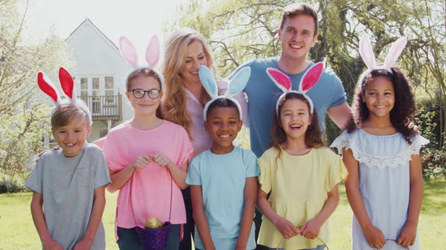 Portrait-of-group-of-parents-and-children-wearing-bunny-ears-on-Easter-egg-hunt-in-garden-smiling-at-camera---shot-in-slow-motion