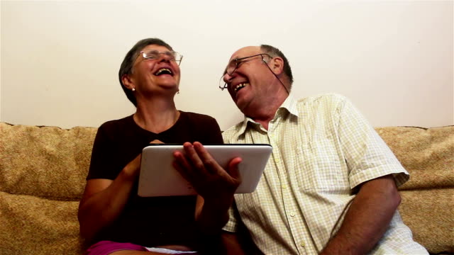 An-elderly-couple-is-sitting-on-a-sofa-at-home-and-watching-photos-on-a-tablet-pc,-smiling-and-talking-actively.