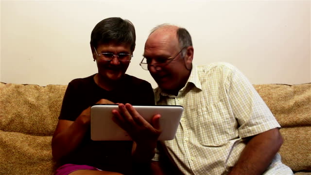 Una-pareja-de-ancianos-está-sentada-en-un-sofá-en-casa-y-viendo-fotos-en-una-tableta-PC,-sonriendo-y-hablando-activamente.