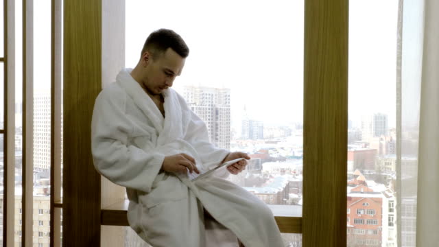 Man-dressed-in-white-bathrobe-sitting-on-windowsill,-using-tablet-computer.-4K