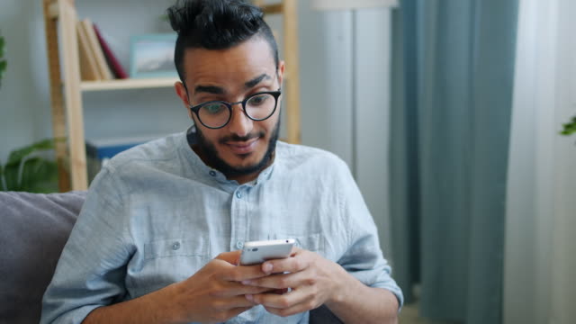 Happy-young-Arab-using-smartphone-looking-at-screen-with-happy-face-at-home