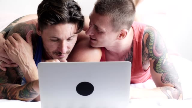 Schwules-Paar-im-Bett-mit-Laptop-Computer.-Küssen-in-Wange.