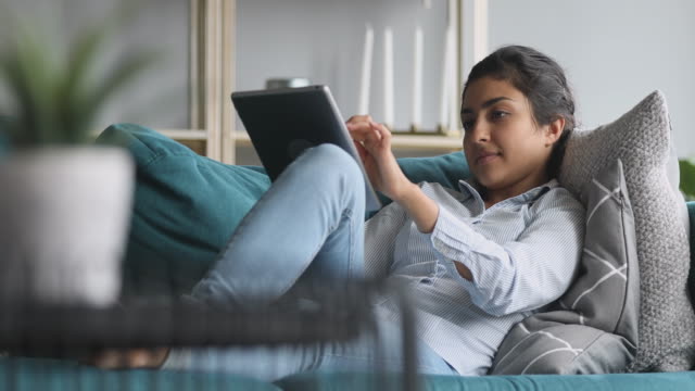 Mujer-india-feliz-relajarse-en-el-sofá-sosteniendo-usando-la-tableta-digital