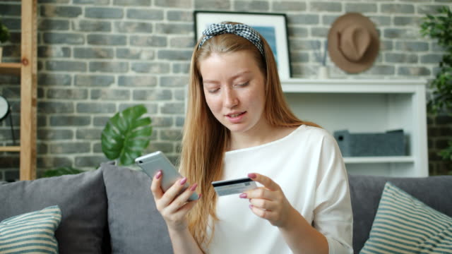 Cheerful-lady-shopping-online-paying-with-credit-card-using-smartphone-at-home