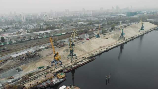 Aerial-view-of-cargo-port-cranes-near-a-pile-of-sand-with-ships,-scows-and-barges-transporting-river-sand.-Industrial-city-covered-with-smog-and-fog