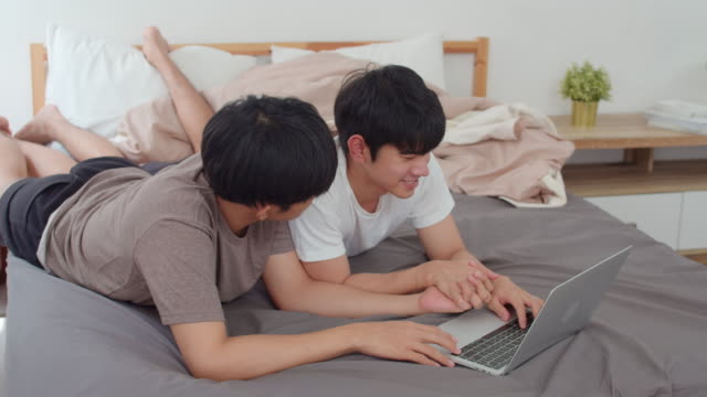 Asiático-Gay-lgbtq-hombres-pareja-usando-computadora-portátil-en-casa-moderna.-Joven-amante-de-Asia-hombre-feliz-descanso-juntos-después-de-despertar,-viendo-película-acostado-en-la-cama-en-la-habitación-en-casa-en-el-concepto-de-la-mañana.