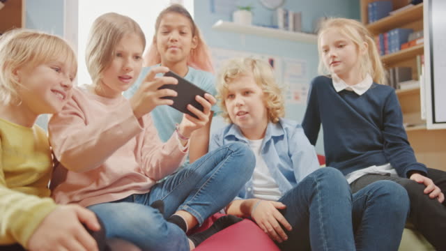 Kinder-Surfen-im-Internet-und-Online-Videospiele-auf-dem-Handy-spielen,-Videos-ansehen.-Diverse-Gruppe-von-niedlichen-kleinen-Kindern-sitzen-zusammen-auf-den-Bean-Bags-verwenden-Smartphone-und-sprechen,-viel-Spaß.