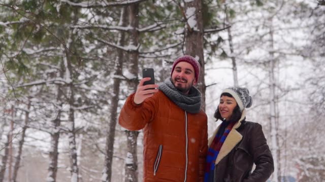 Joven-pareja-hipster-amante-tomando-selfie-foto-bajo-nevada-en-el-bosque.-Bloger-hombre-disparando-en-la-cámara-del-teléfono-inteligente-con-novia-para-las-redes-sociales.-Familia-Influencer-disfrutando-de-vacaciones-de-Navidad