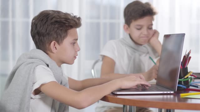 Portrait-of-Caucasian-schoolboy-looking-at-his-twin-brother-playing-games-on-laptop-and-sighing.-Brunette-boy-gesturing-yes-and-typing-again.-Different-lifestyles,-internet-addiction,-social-media.
