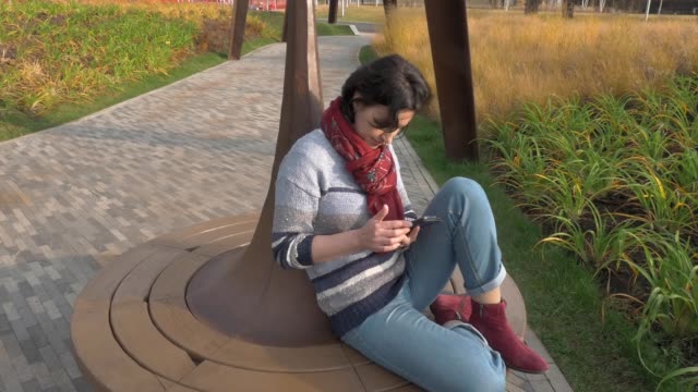 Mujer-caucásica-sentada-en-un-banco-redondo-del-parque-con-un-teléfono-en-las-manos