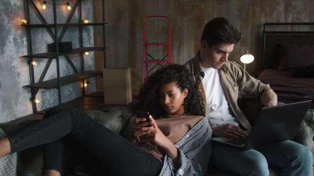 Couple-moving-to-new-apartment.-He-working-at-laptop,-she-using-smartphone.-Relaxing-together-on-gray-couch-in-room-with-cardboard-boxes.-Close-up