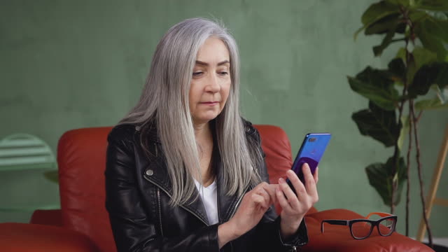 Happy-smiling-mature-woman-with-long-gray-hair-sitting-in-comfortable-chair-and-using-her-phone