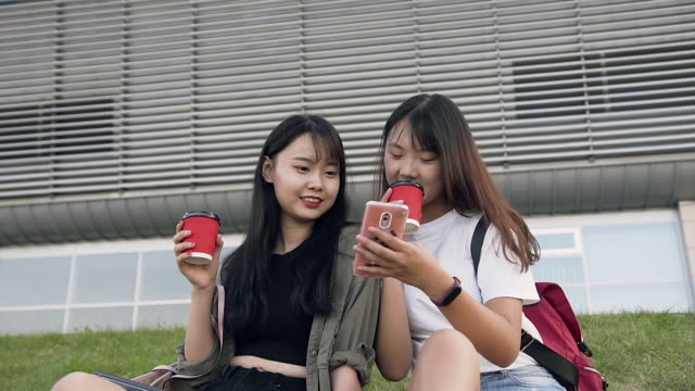 Attraktives-Porträt-von-zwei-angenehm-fröhlichen-asiatischen-Brünetten,-die-auf-Gras-sitzen-und-mit-ihren-Handys,-Kaffee-trinken-auf-dem-modernen-Gebäude-Hintergrund