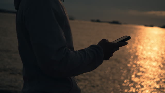 Close-up-athlete-runner-using-smartphone-typing-message-during-resting-after-running-exercising-outdoors-on-the-beach-at-beautiful-sunset-in-summer.