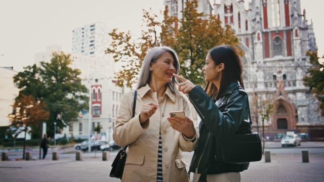 Asiatische-reife-Mutter-und-erwachsene-Tochter-verwenden-Smartphone,-während-sie-auf-dem-Stadtplatz-stehen,-lächelnd-und-auf-etwas-zeigen