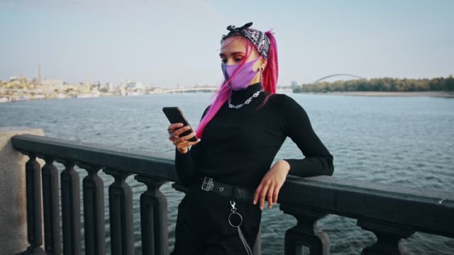 Hipster-Frau-in-informellen-Outfit-und-Schutzmaske-ist-mit-ihrem-Handy,-während-auf-Zaun-der-Stadt-Damm-lehnen