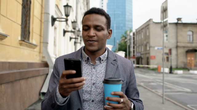 Steadicam-shot-of-young-happy-businessman-using-smartphone-and-walking-with-cup-of-coffee-outdoors