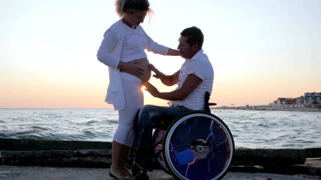 invalid-spouse-in-wheelchair-with-pregnant-female-at-sunset,-handicapped-man-in-wheelchair-caresses-woman