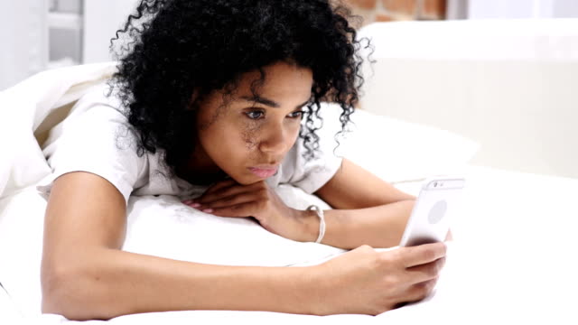 Cansado-mujer-Afro-Americana-en-la-cama-de-desplazamiento-y-navegación-en-Smartphone