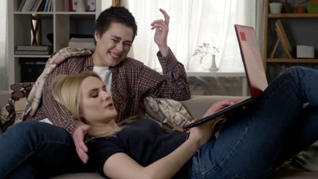 Young-blond-woman-lies-on-the-lap-of-a-brunette-on-the-couch-and-using-red-laptop,-cosiness,-cuddles,-plaid,-talking,-lesbians,-lgbt,-discussion-60-fps