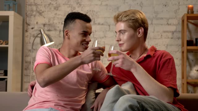 Pareja-gay-multinacional-sentado-en-un-sofá,-tintinear-de-vasos-y-bebiendo-vino.-Los-amantes-de-la-LGBT,-muchachos,-jóvenes-amigos,-feliz-concepto-de-familia-gay.-60-fps