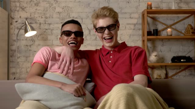 Two-gay-guys-are-sitting-on-a-sofa-covered-in-a-warm-blanket-and-watching-a-3d-funny-film,-they-are-happy-and-laughing.-LGBT-lovers,-happy-gay-family,-home-cosiness-concept.-Look-at-the-camera-60-fps
