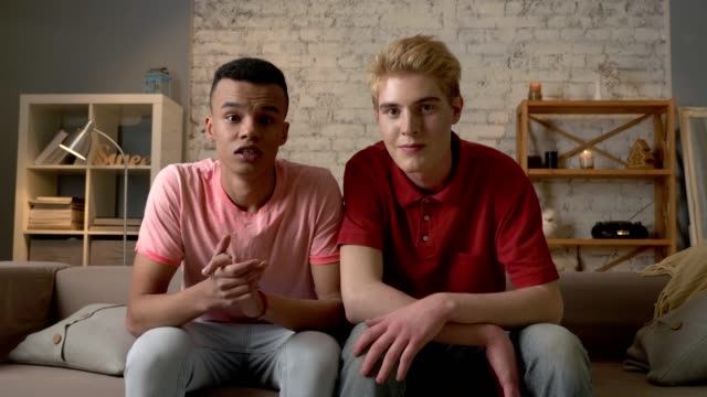Two-gay-guys-are-sitting-on-the-couch-and-watching-TV,-emotion-of-expectation-and-rejoice,-laughing.-LGBT-lovers,-happy-gay-family,-home-cosiness-concept.-Look-at-the-camera-60-fps