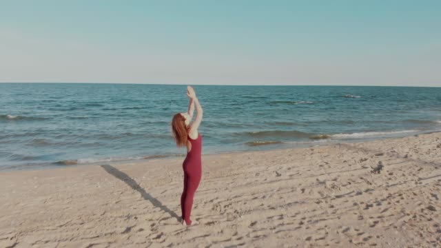 Drone-bewegt-sich-hinter-Frau-in-Sport-Wear-hält-Yoga-Asana-Position-am-Meer-oder-Ozean-Sandstrand.-Windige-sonniges-Wetter.-Luftaufnahme-des-friedlichen-Gesundheit-Mädchen-Praxis-durchführen