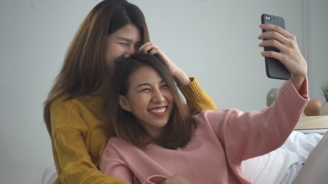 Beautiful-young-asian-women-LGBT-lesbian-happy-couple-sitting-on-sofa-hug-and-using-phone-taking-selfie-together-bedroom-at-home.-LGBT-lesbian-couple-together-indoors-concept.-Spending-nice-time-home.