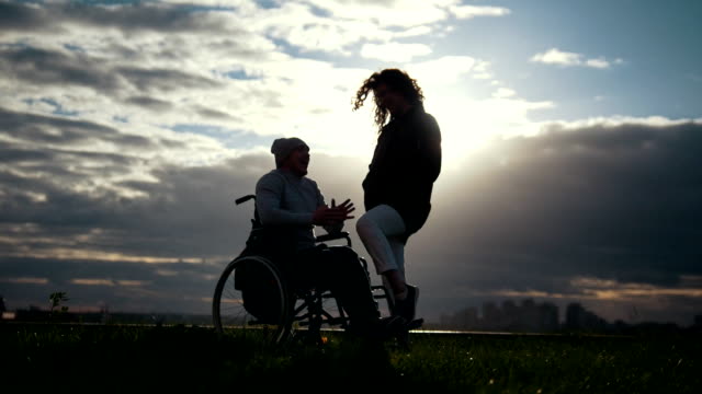 Pflege-von-Menschen-mit-Behinderungen---Mann-im-Rollstuhl-mit-Frau-sprechen-in-den-bewölkten-Sonnenuntergang