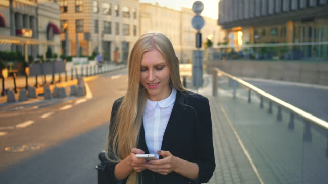 Formal-business-woman-walking-on-street.-Elegant-blond-woman-in-suit-and-walking-on-street-and-browsing-smartphone-with-smile-against-urban-background