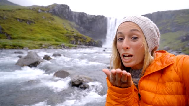 Junge-Frau-unter-Selfie-Porträt-mit-herrlichen-Wasserfall-in-Island,-weht-einen-Kuss-auf-Kamera.-Die-Leute-reisen-Exploration-Konzept