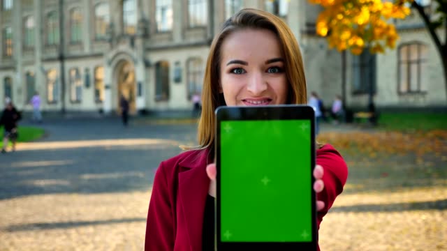 Young-attractive-female,-student-showing-screen-of-the-tablet-to-camera,-outside-the-university