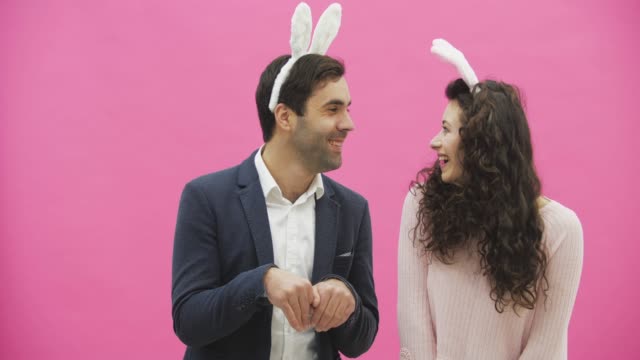 Beautiful-young-couple-standing-on-pink-background.-At-the-same-time,-they-smile-to-recreate-the-movements-of-hares-after-kissing.-Together-with-the-pink-paws-on-the-head.-Happy-family-is-preparing-for-Easter.