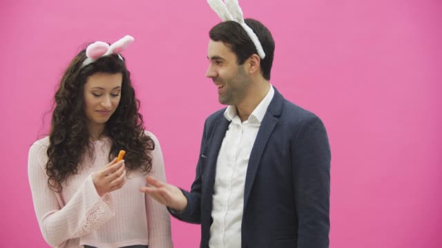 Young-couple-are-beautiful-on-pink-background.-During-this-time,-they-are-dressed-in-rabble-ears.-Looking-at-each-other.-A-man-shows-carrots-and-his-wife-starts-to-laugh.-A-young-man-begins-to-choke-the-girl.