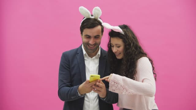 Young-lovers-couple-on-pink-background.-With-ravenous-ears-on-the-head.-During-this-Easter-photo,-sephi-do-on-the-phone-and-looking-at-them-rejoice.