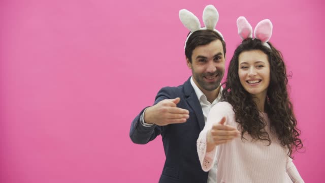 Young-creative-couple-on-pink-background.-With-hackneyed-ears-on-the-head.-During-this-time,-they-show-the-gestures-of-the-call-and-look-at-the-other-side-together.