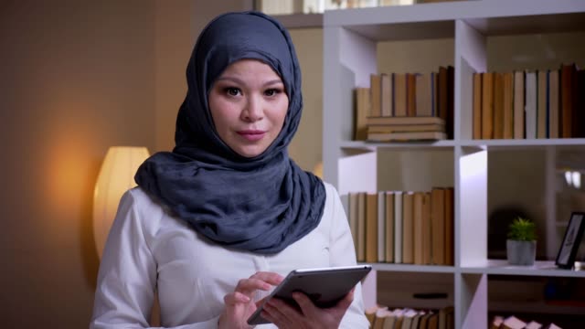 Closeup-shoot-of-adult-muslim-businesswoman-using-the-tablet-looking-at-camera-and-smiling-happily-on-the-workplace-indoors
