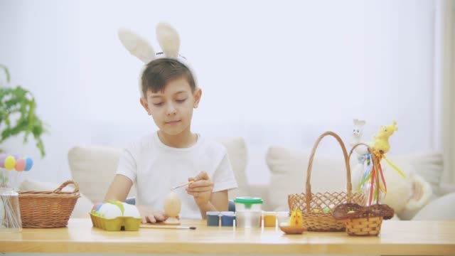 El-chico-creativo-está-coloreando-un-huevo-de-Pascua-con-la-ayuda-de-pincel-pintura.,-usando-orejas-de-conejo,-luego-levanta-los-dedos-del-pulgar-hacia-arriba.