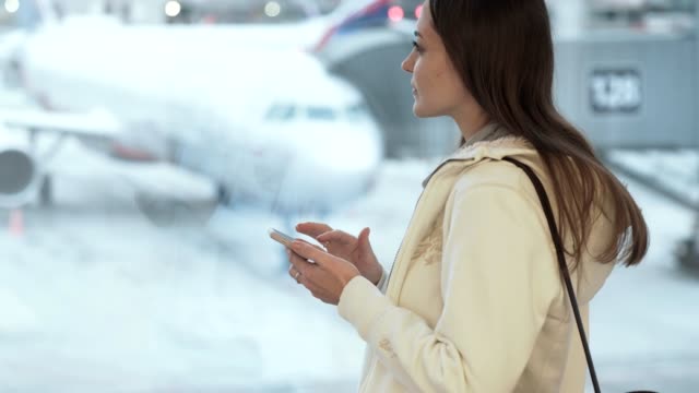 Side-view-beautiful-girl-with-long-hair-standing-at-window-in-airport-and-texting-in-phone-on-background-of-airplane
