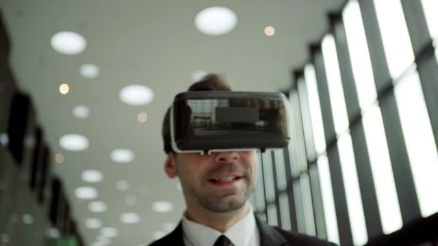 Close-up-shot-of-middle-aged-businessman-in-virtual-reality-headset-having-video-call-and-talking-while-walking-along-office-corridor