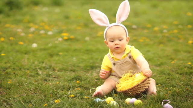 Schönes-Baby-in-einem-Osterhasen-Kostüm-sammelt-Ostereier-in-einem-Korb,-der-auf-dem-Rasen-im-Park-sitzt.-Frühlingshaftes,-glückliches-Osterfest