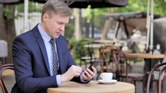 Businessman-Upset-by-Loss-on-Smartphone,-Sitting-in-Outdoor-Cafe