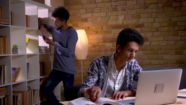 Closeup-shoot-of-two-culturally-diverse-students-learning-in-the-school-library-indoors.-Indian-male-studying-online-on-the-laptop-while-african-american-man-using-the-tablet-on-the-background