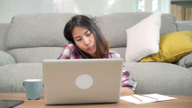 Freelance-Asian-woman-working-at-home,-business-female-working-on-laptop-and-using-mobile-phone-talking-with-customer-on-sofa-in-living-room-at-home.-Lifestyle-women-working-at-home-concept.