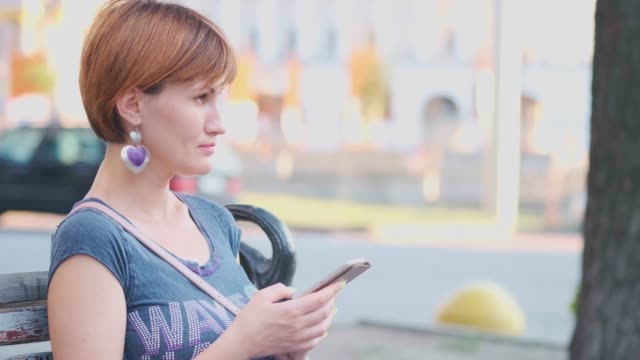 Woman-Using-Smartphone-Relaxes-on-the-Bench-t-the-street.-SLOW-MOTION,-4K.-Young-Millennial-Woman-in-Arboretum-making-gestures-on-Phone-Display.