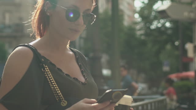 Young-attractive-girl-writing-a-text-message-on-her-smartphone-at-the-subway-exit-in-street,-holding-her-coffee,-during-sunny-summer-in-Paris.-Trendy-and-cool,-tilt-down.-Freckles,-sunglasses.