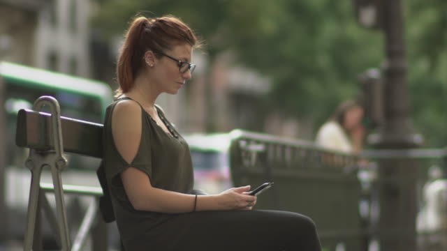 Attractive-redhead-woman-with-glasses,-freckles,-piercings-and-red-hair-writing-a-text-message-on-her-smartphone-sitting-on-street-bench,-during-sunny-summer-in-Paris.-Slow-motion.-Trendy.